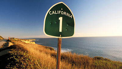 A green highway sign that says California 1 with ocean and coastline in background
